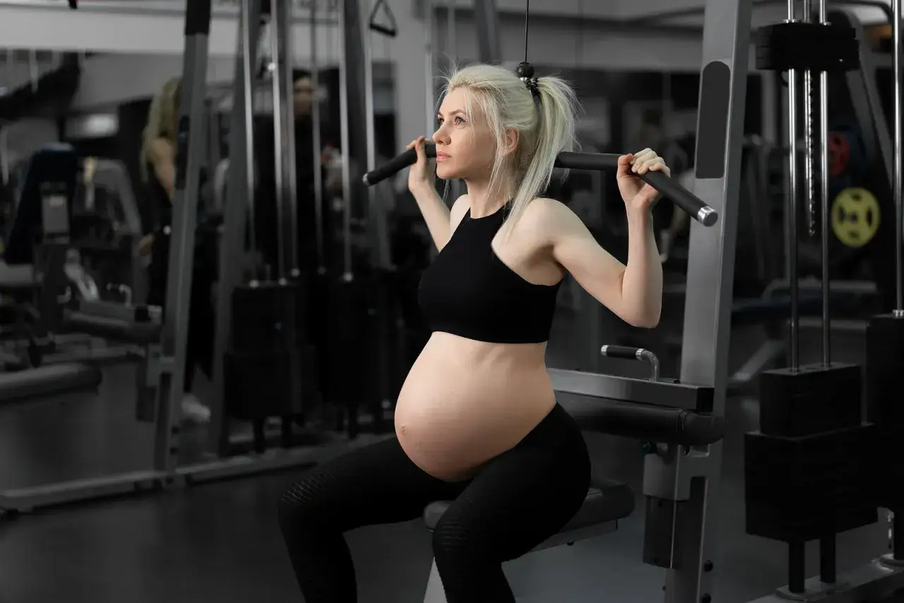 Gravid kvinna tränar marklyft i gym. Hon sitter på en träningsmaskin och drar i en stång.
