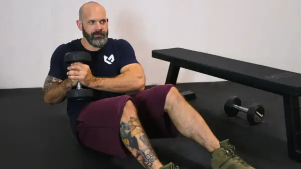 Bald man with beard doing Russian twists with a dumbbell, sitting on a gym floor.