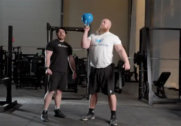 Man med skägg utför en kettlebell press med en blå kettlebell. En annan man står bredvid och tittar.